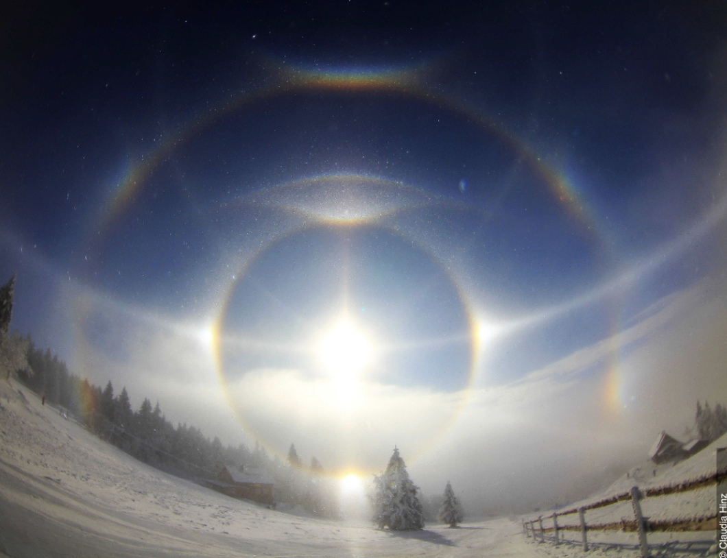 Cover image from Claudio Hinz depicting a complex halo containing a 22-degree halo, parhelic circle, sundogs, lower and upper tangent arcs (with the upper one having a suncave parry arc), infralateral arc, heliac arc, a supralateral arc touching a circumzenithal arc, and 4 tape arcs.
